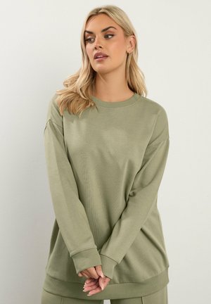 Vrouw met blond haar draagt een oversized olijfgroene trui, staat met gevouwen handen en kijkt opzij tegen een effen achtergrond.