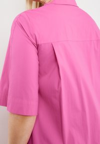 Chemise rose à manches courtes avec une coupe décontractée, dotée d'un col pointu et d'une texture subtile. Un pli dans le dos améliore le mouvement.