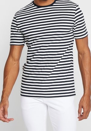 Mężczyzna w czarno-białej poziomej paskowanej koszulce z krótkim rękawem i białych spodniach na jednolitym tle.