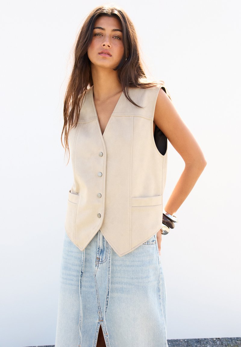 Beige ärmellose Weste mit V-Ausschnitt, vorderem Knopfverschluss und zwei Taschen, kombiniert mit einem hellblauen Jeansrock mit einem Schlitz vorne.