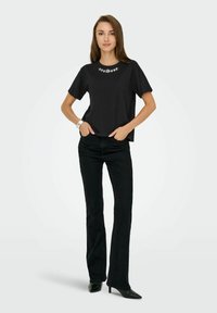 Zwarte T-shirt met korte mouwen, voorzien van witte bloemige borduursels bij de halslijn, gecombineerd met zwarte flared jeans en puntige zwarte hakken.