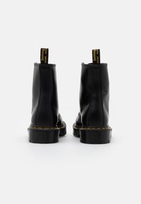 Bottines en cuir noir avec une texture lisse, couture jaune le long de la semelle et une languette pour un enfilage facile. Semelle plate et robuste.