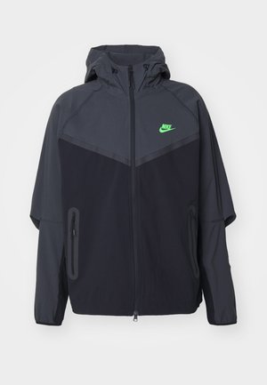 Chaqueta ligera en negro y gris oscuro con capucha, que presenta un logo de Nike en verde, bolsillos con cremallera y un diseño aerodinámico y deportivo.