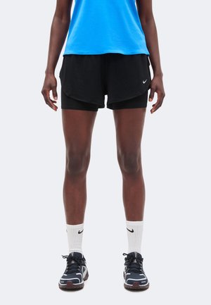 Person iført sorte lag-på-lag løbeshorts, hvide Nike-sokker, sorte og hvide sneakers og en blå top, stående med benene lige.