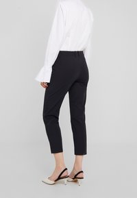 Černé kalhoty na míru se slim fit střihem, zkrácenou délkou ke kotníkům a hladkým povrchem, kombinované s bílou blůzou a nazouvacími podpatky.