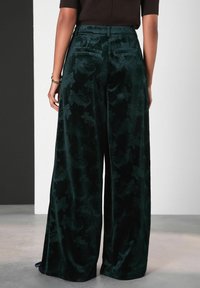 Persoană îmbrăcată în pantaloni largi, cu talie înaltă, din catifea verde închis, cu modele florale subtile, stând pe un podea gri, în fața unui fundal alb și negru.