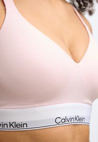 Απαλό ροζ μπραλέτο με σχεδίαση βαθιάς V λαιμόκοψης και λεία υφή. Διαθέτει λευκή ταινία με μαύρη επιγραφή "Calvin Klein".