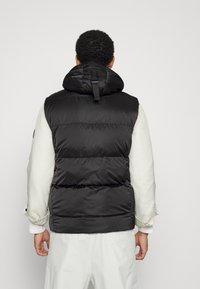 Mężczyzna ubrany w czarną, ocieplaną kamizelkę bez rękawów z kapturem, pod którą ma na sobie białą koszulę z długim rękawem oraz białe spodnie, stoi tyłem na jednolitym tle.