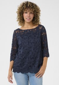 Top in pizzo blu navy con scollatura a barca, maniche a tre quarti, orlo scallop e motivo floreale. Indossato con jeans blu chiaro.