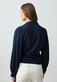 Blusa blu navy con maniche lunghe e voluminose, colletto e design aderente. Tessuto liscias con polsini abbottonati e silhouette sartoriale.