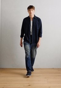Chemise bleu marine à boutons, débardeur blanc, jean bleu délavé et baskets sombres sur un modèle marchant sur un sol en bois contre un mur clair.