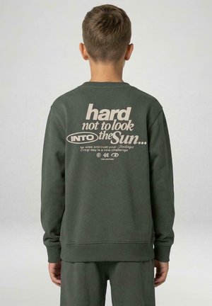 Jongen met kort haar die een donkergroene trui draagt met tekst "hard not to look into the sun", staand voor een effen lichte achtergrond.