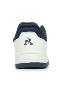 Sneaker bianca con finitura in pelle liscia, collarino in tessuto blu navy, suola testurizzata e logo nero sul tallone. Presenta un design low-top.