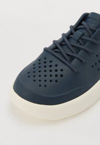 Tumedat sinised sneakersid tekstureeritud pinnaga, ventilatsiooniavadega hingavuse tagamiseks, lamedate paeltega ja valgest kummist tallaga.