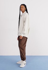 Υπερμεγέθης λευκή μπλούζα με γιακά, συνδυασμένη με καφέ παντελόνι και λευκά αθλητικά παπούτσια με γκρίζες λεπτομέρειες. Απαλή υφή και άνετη εφαρμογή.
