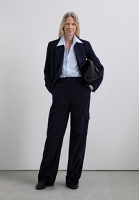 Blazer de pana en color navy y pantalones cargo a juego, combinados con una camisa de color azul claro y un bolso negro. Textura suave y corte relajado.