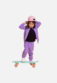 Dziecko na jasnoniebieskim deskorolce, ubrane w fioletowy dres i różową czapkę. Deskorolka ma różowe kółka i fakturowaną powierzchnię.