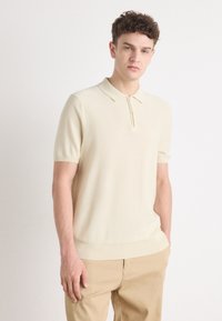 Kortärmad polotröja i ljusbeige stickad tyg, med en dragkedja i kragen och ribbad fåll. Bärs tillsammans med beiga byxor.