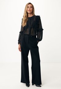 Blusa nera trasparente con maniche a sbuffo, rouches e motivo a pois; pantaloni neri a gamba larga; scarpe nere con tacco; dettagli minimal con bottoni.