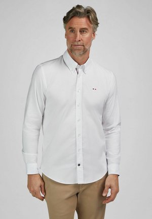 Man met grijs haar en baard, gekleed in een wit overhemd en beigekleurige broek, staand tegen een effen lichtgrijze achtergrond.