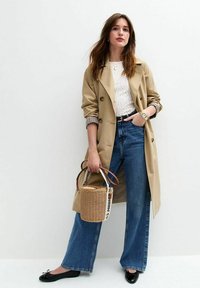 Beige trenchcoat met grote knopen, blauwe high-waisted jeans, zwarte platte schoenen en een geweven ronde handtas met een gestreepte schouderband.