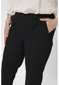 Persona vestida con pantalones negros y una blusa blanca con un patrón texturizado, una mano descansando en el bolsillo y la otra a su lado con pulseras de perlas.