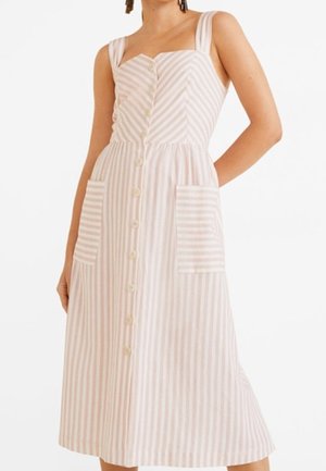 Vrouw die mouwloze jurk tot op de knie draagt met verticale roze en witte strepen, knopen aan de voorkant en grote voorzakken.