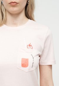 Γυναίκα που φοράει ροζ ανοιχτό t-shirt με εκτύπωση τσέπης που δείχνει ένα βάζο με λευκό smoothie φράουλας και δύο φράουλες, με την επιγραφή «Smoothie Φράουλα».