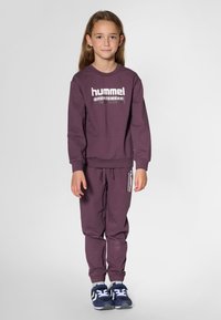 Lila Baumwoll-Sweatshirt mit "hummel"-Logo, kombiniert mit passenden Jogginghosen mit Logoakzenten. Dunkle Sportschuhe vervollständigen das Outfit.