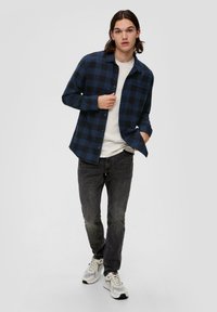 Marineblauw en zwart geruit flanellen shirt met knoopsluiting, gedragen over een witte t-shirt, gecombineerd met donkere spijkerbroek en sportschoenen.