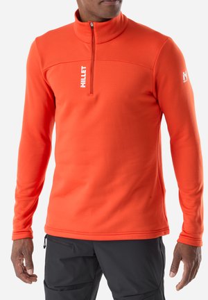 Man wearing bright orange half-zip pullover with white "Millet" logos, paired with black pants, standing against white background.