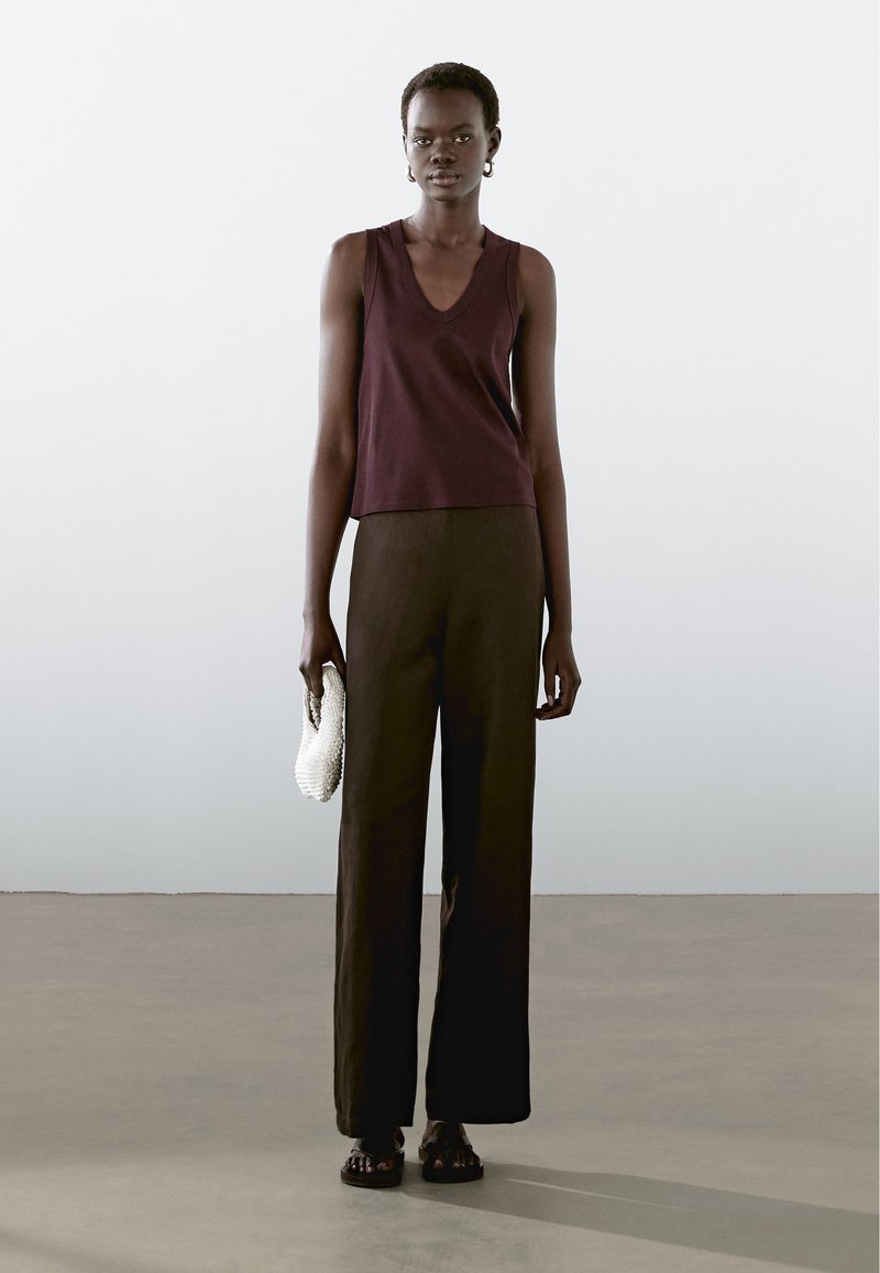 Top bordeaux sans manches avec un col en V, associé à un pantalon noir large taille haute et une petite pochette blanche.