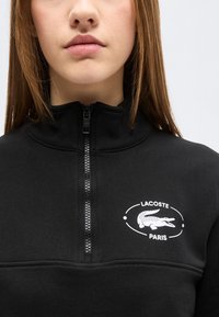 Νέος που φοράει μαύρο φούτερ Lacoste Paris με φερμουάρ και το λευκό λογότυπο κροκοδείλου στο στήθος, σε απλό φωτεινό φόντο.