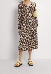 Černé květované šaty s dlouhým rukávem, výstřihem do V a staženým pasem, zdobené výraznými oranžovými a růžovými květy. Doplněné o robustní sandály s volánky.