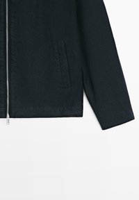 Manche de veste noire et section inférieure du torse avec fermeture éclair verticale et une seule poche sur fond blanc uni.