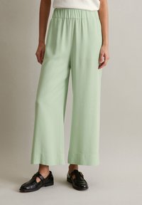 Helle grüne weit geschnittene Hose mit lockerer Passform, elastischem Bund, glatter Stofftextur und sauberem Saum. Getragen mit schwarzen Slippern.
