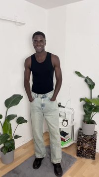 Camisola preta sem mangas, jeans largos azul claro, botas de couro pretas. Ambiente interior com plantas verdes e uma máquina de costura ao fundo.