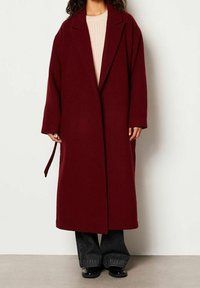 Femme portant un long manteau bordeaux, un pull rose clair, un jean large foncé et des chaussures noires sur un fond clair uni.