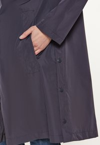 Giacca impermeabile di un viola scuro con tasca laterale, dettagli con bottoni e una texture liscia. Le maniche sono leggermente ampie.