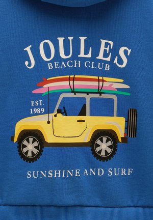 Gelbes Geländefahrzeug mit vier Surfboards auf dem Dach, Text "Joules Beach Club", "Est. 1989" und "Sunshine and Surf" auf blauem Stoff.