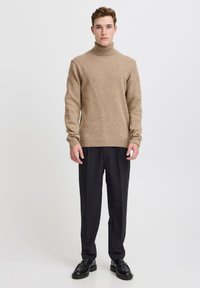 Maglione a collo alto beige in tessuto morbido, indossato con pantaloni sartoriali grigio scuro a quadri e scarpe nere, in piedi davanti a uno sfondo bianco.