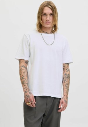 Witte katoenen t-shirt met korte mouwen en een rechte snit. Kenmerkt zich door een ronde hals en zichtbare tatoeages op de armen. G gedragen met grijze broeken.