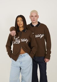Cardigan zippé en tricot marron avec le texte "Legacy" en blanc, présentant un col noir avec des rayures blanches. Modèle porté par deux personnes.