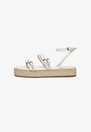 Hvid flad sandal med fletet rebssål, tre tynde remme med sølvspænder og en ankelrem med spænde lukning på hvid baggrund.
