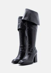 Bottes hautes jusqu'aux genoux en cuir noir avec revers rabattus, fermetures éclair latérales, bouts ronds et talons blocs sur fond blanc.