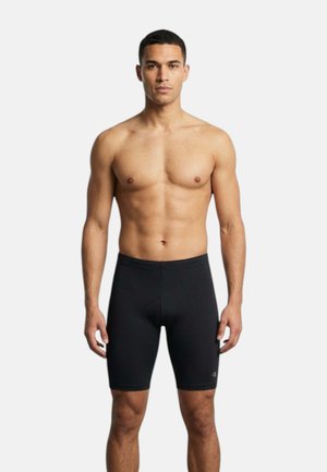 Uomo atletico in piedi di fronte, indossa pantaloncini da ciclismo neri aderenti, su sfondo bianco semplice.