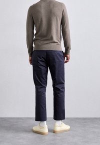 Bruine gebreide trui, marineblauwe broek, witte sokken, gele sneakers. Achteraanzicht toont de pasvorm van de trui, de lengte van de broek en de stijl van de schoenen.