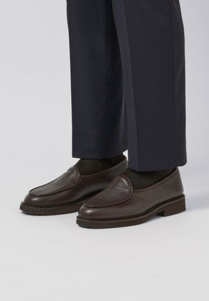 Hnědé kožené nazouvací boty s texturovaným povrchem, kulatou špičkou a nízkou gumovou podrážkou, kombinované s tmavými kalhotami a ponožkami.