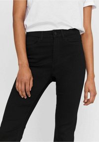 Personne portant un jean skinny noir taille haute et un t-shirt blanc à manches courtes, debout les mains détendues le long du corps.