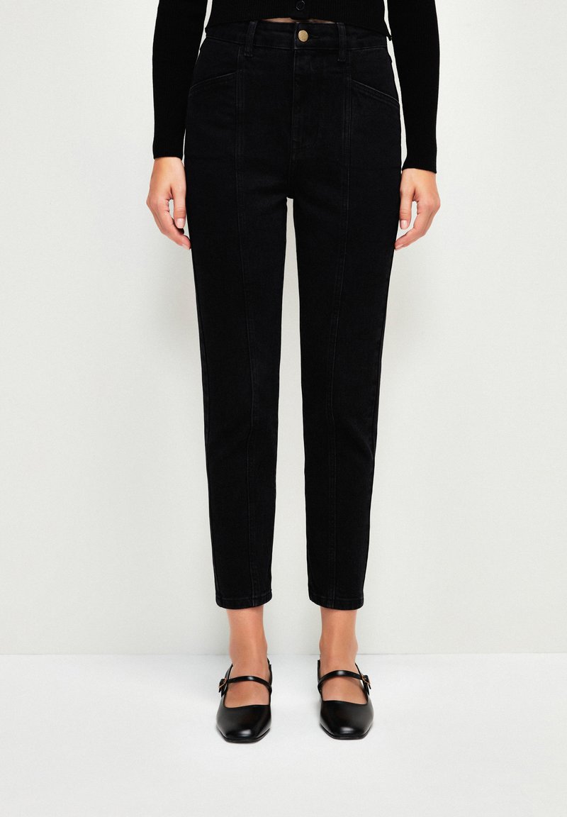 Jeans negros de cintura alta con ajuste tapered, tela de denim texturizada, bolsillos frontales y cierre de botón dorado. Se incluye un par de ballerinas negras.
